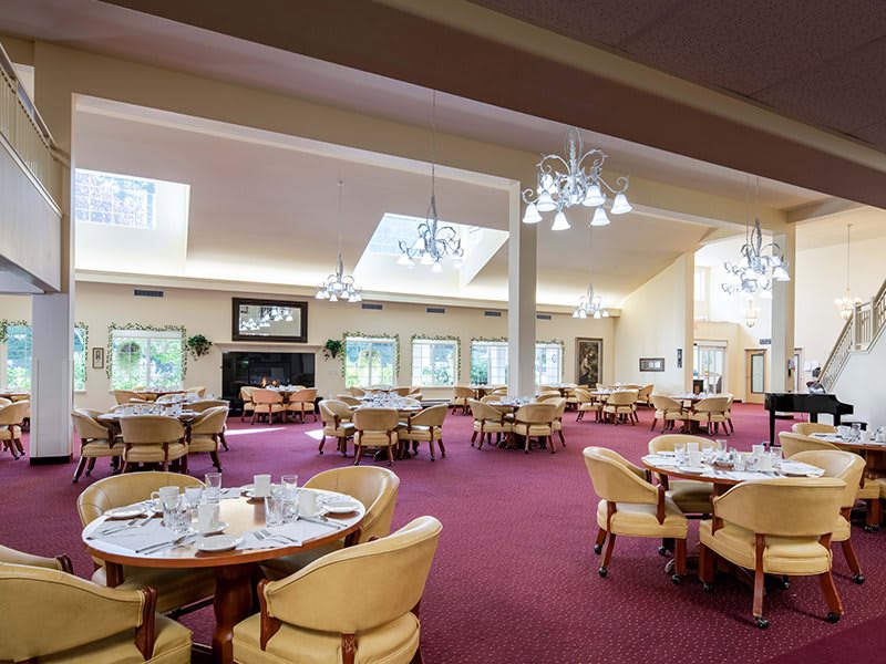 A spacious and well-lit dining area with multiple tables and chairs, featuring elegant chandeliers and a rich carpeted floor.
