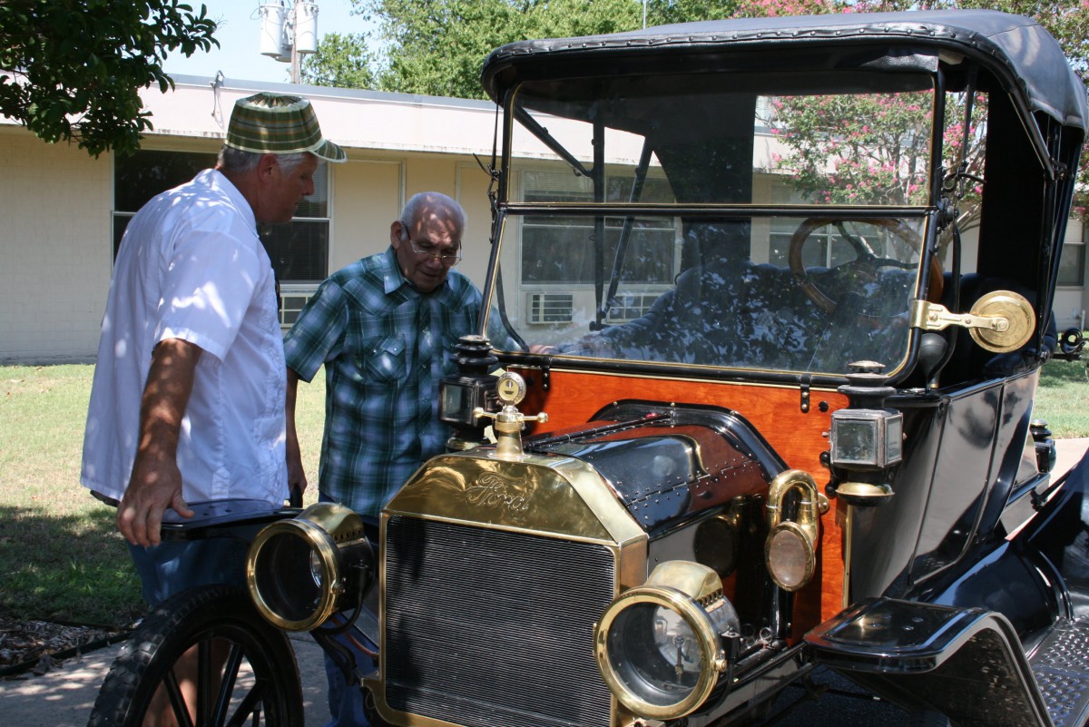 Two individuals admire a vintage car displayed outdoors.