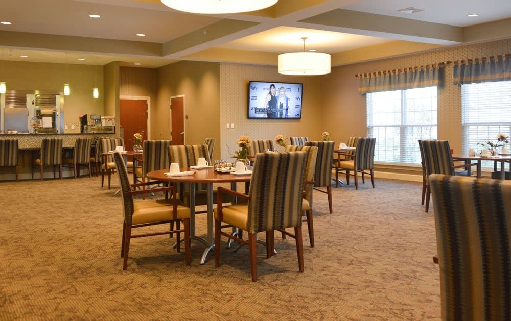 A spacious dining area with neatly arranged tables and chairs, illuminated by overhead lights, featuring a television on the wall.
