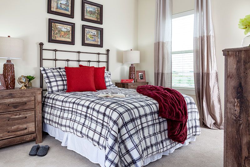 A cozy bedroom featuring plaid bedding with red accent pillows and throw, complemented by framed wall art and soft natural lighting.