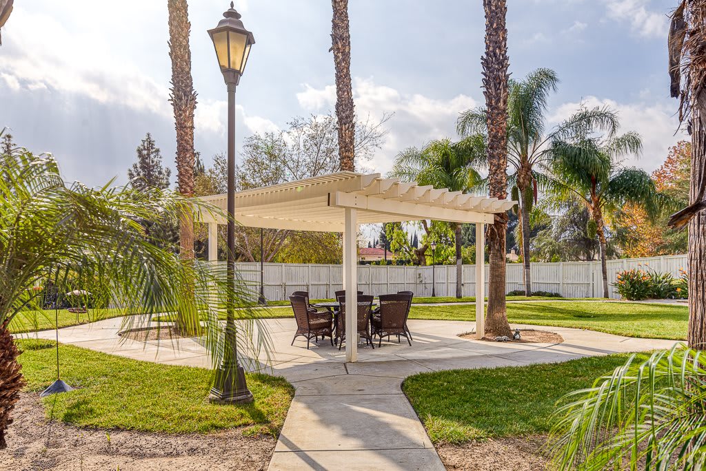 A serene garden setting featuring a gazebo with patio furniture, surrounded by palm trees and lush landscaping.