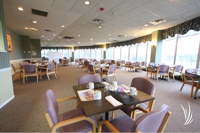 An empty dining hall with neatly arranged tables and chairs, featuring large windows.