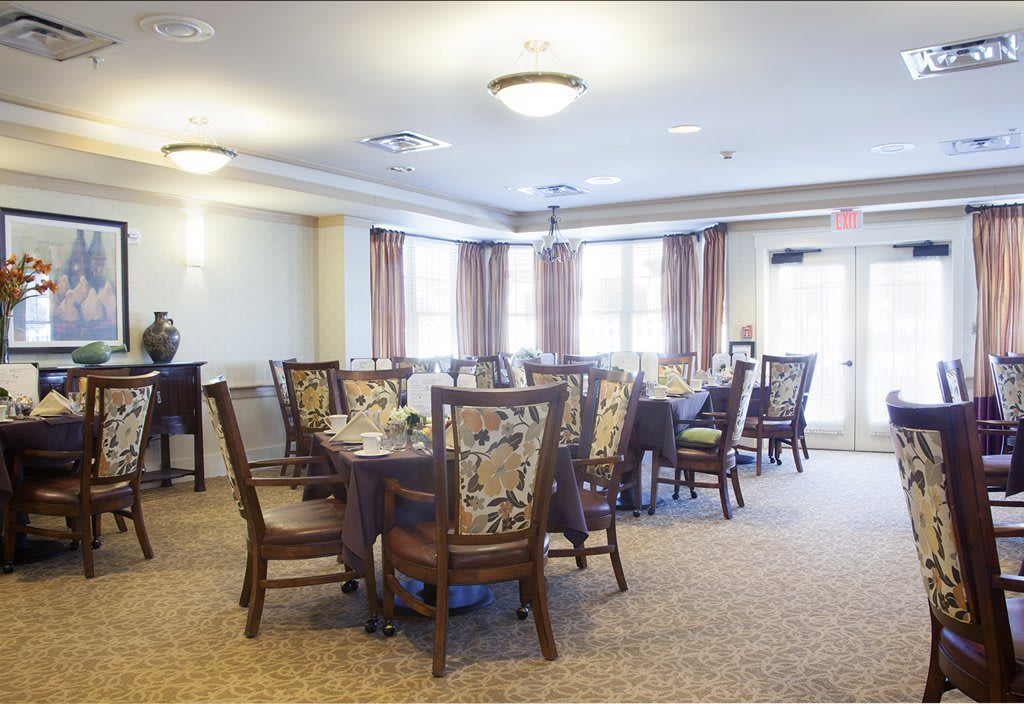 A well-lit dining room with multiple tables and chairs, featuring elegant curtains and a carpeted floor.