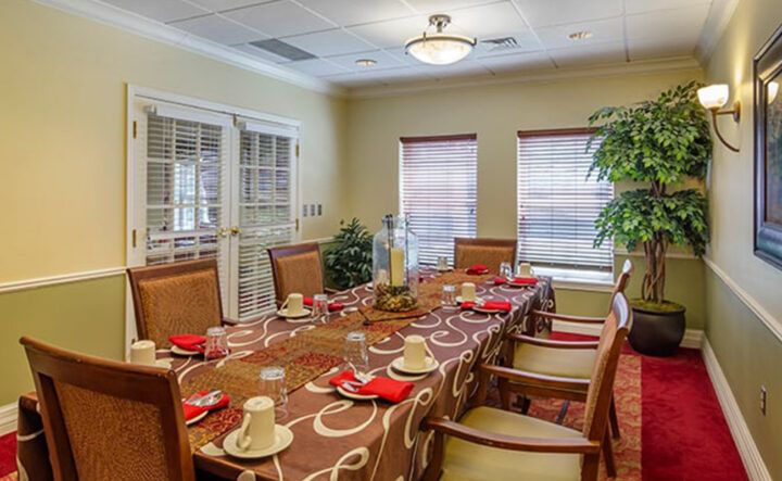 A well-set dining room table with a patterned tablecloth and red napkins, surrounded by wooden chairs, in a warmly lit room.