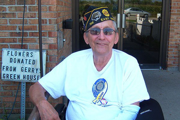 A smiling veteran wearing a hat and glasses is sitting outdoors.