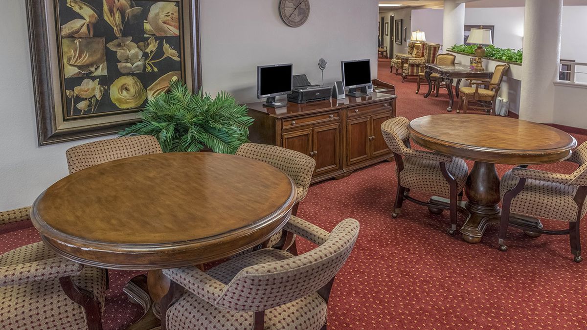A cozy interior space featuring round wooden tables with chairs, computers on a wooden console, and a red carpet.