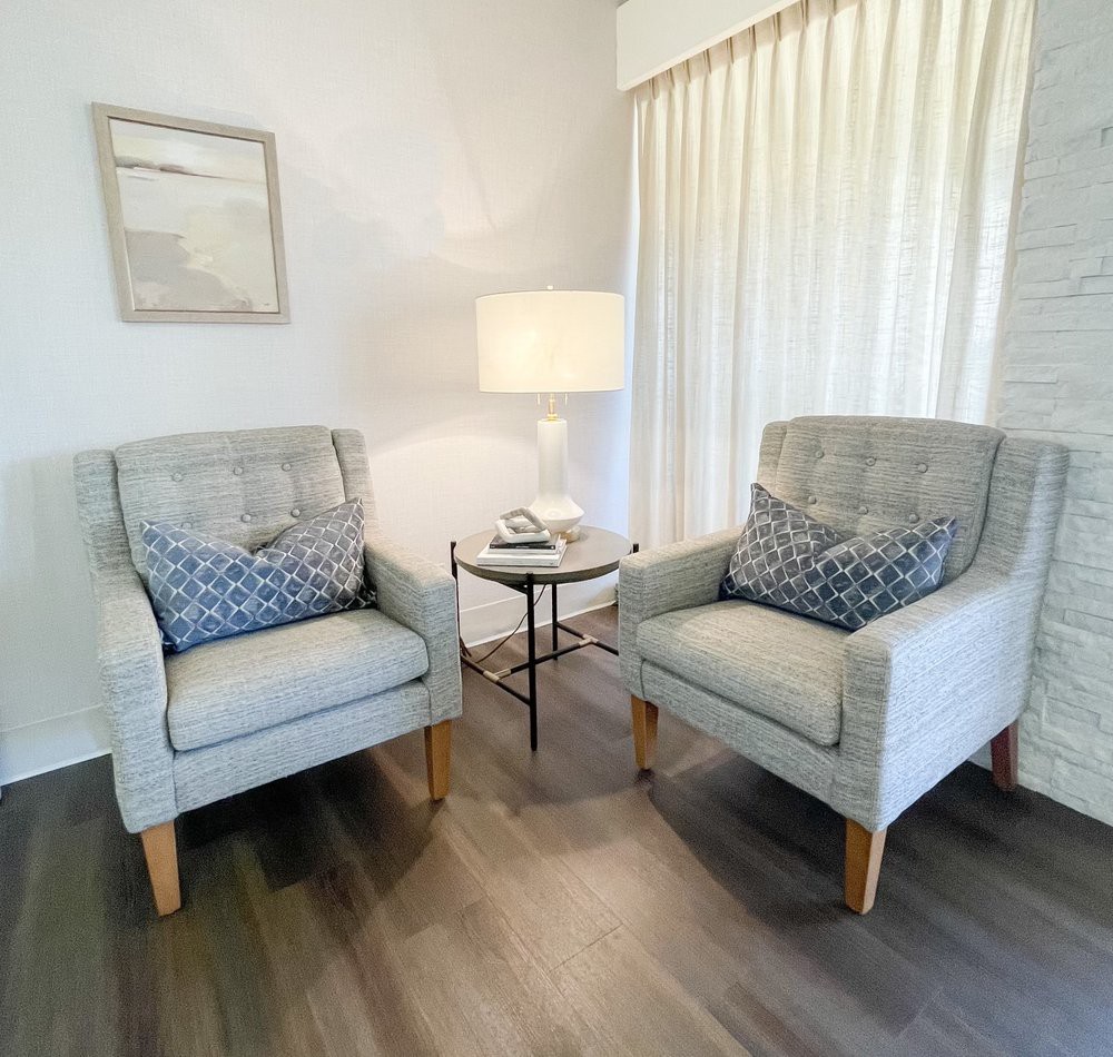 A cozy living room corner featuring two upholstered chairs, a small round table, and a warm lamp.