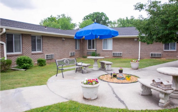 A cozy outdoor patio area featuring a bench, stone table with chairs, and a blue umbrella surrounded by potted plants.