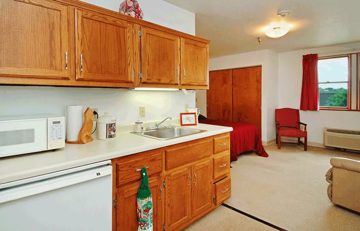A cozy apartment kitchen with wooden cabinets and a sink, leading to a bedroom area.