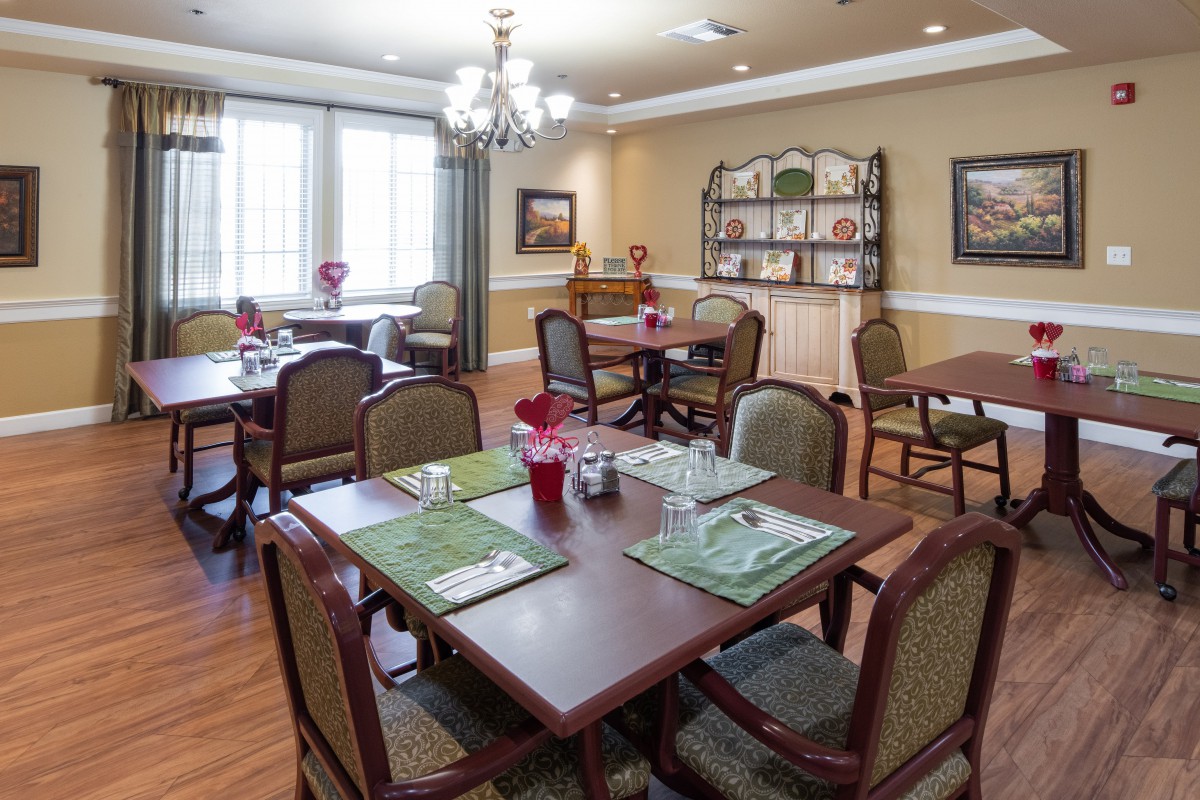 A well-lit dining room with several tables and chairs, decorated with placemats and a chandelier overhead.