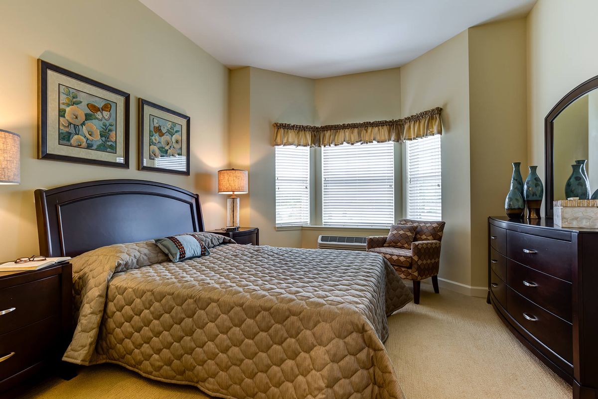 A cozy bedroom with a large bed, two chairs by the window, and warm lighting.