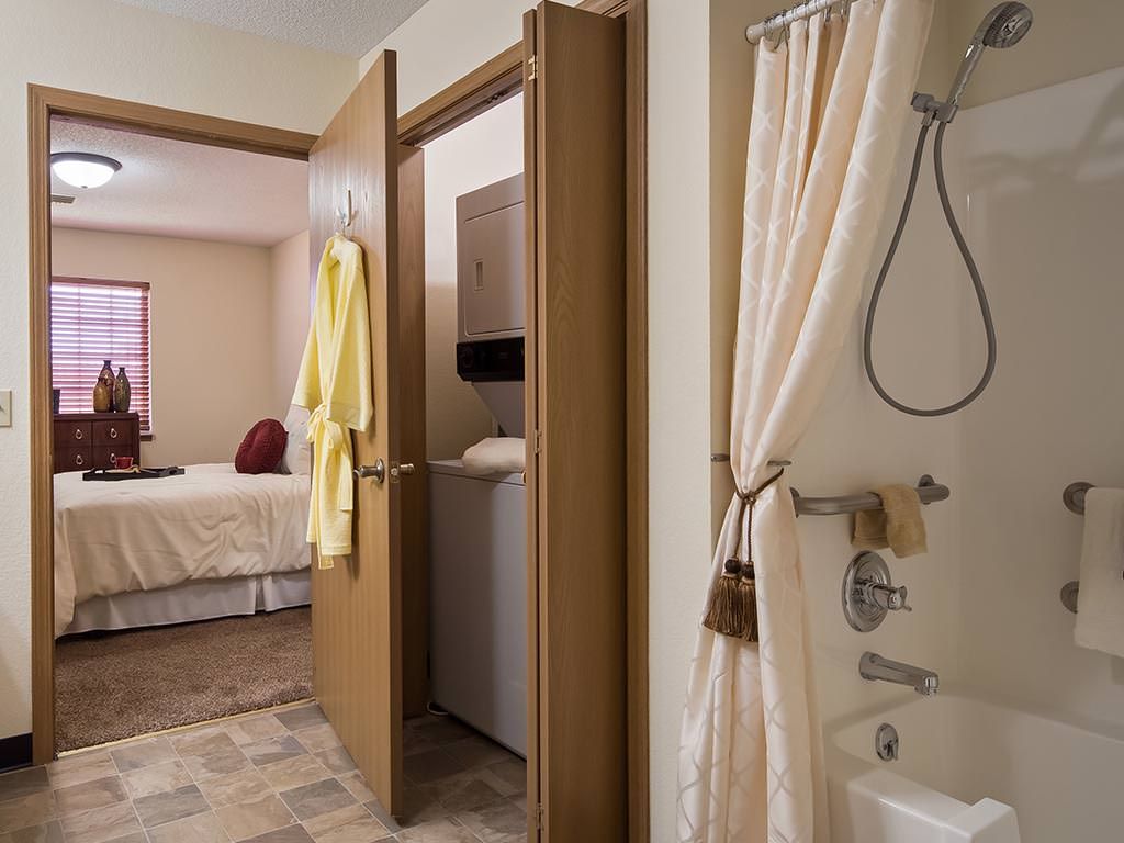 A view from a bathroom showing a shower with a curtain, towels hanging on doors, and an adjacent bedroom.