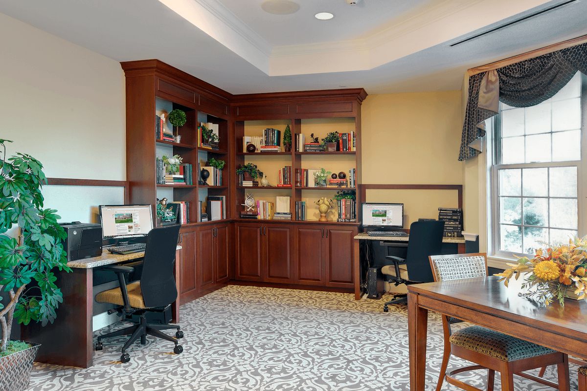 A cozy office space with wooden bookshelves, multiple desks equipped with computers, and comfortable chairs on a patterned carpet.