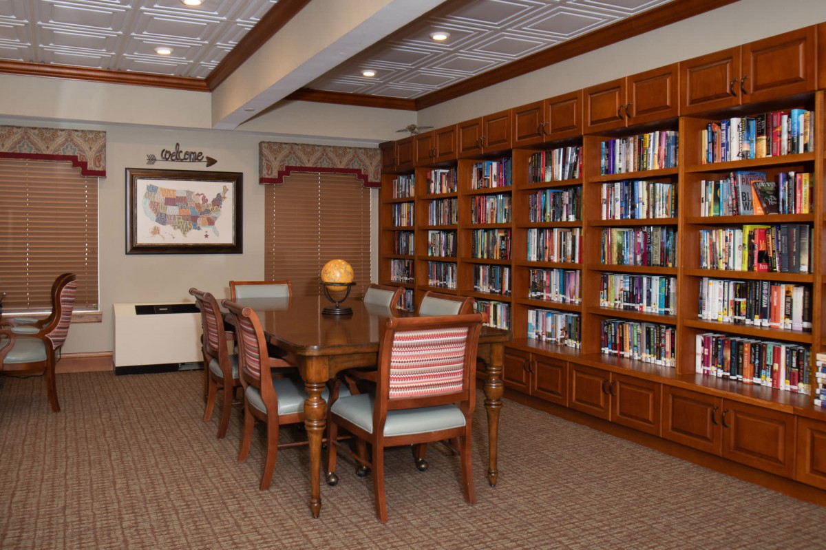 A cozy library room with wooden bookshelves filled with books and a central table surrounded by chairs.
