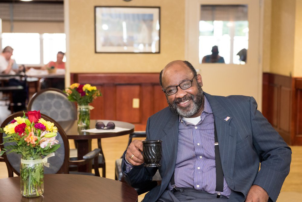 A man sitting indoors, smiling while holding a coffee cup.