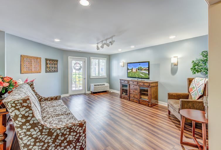 A cozy living room with laminate flooring, featuring a sofa, armchair, and a television mounted on an entertainment center.