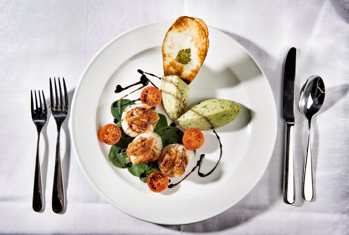 A beautifully plated gourmet dish featuring scallops, garnished with spinach, cherry tomatoes, and a toasted bread slice.