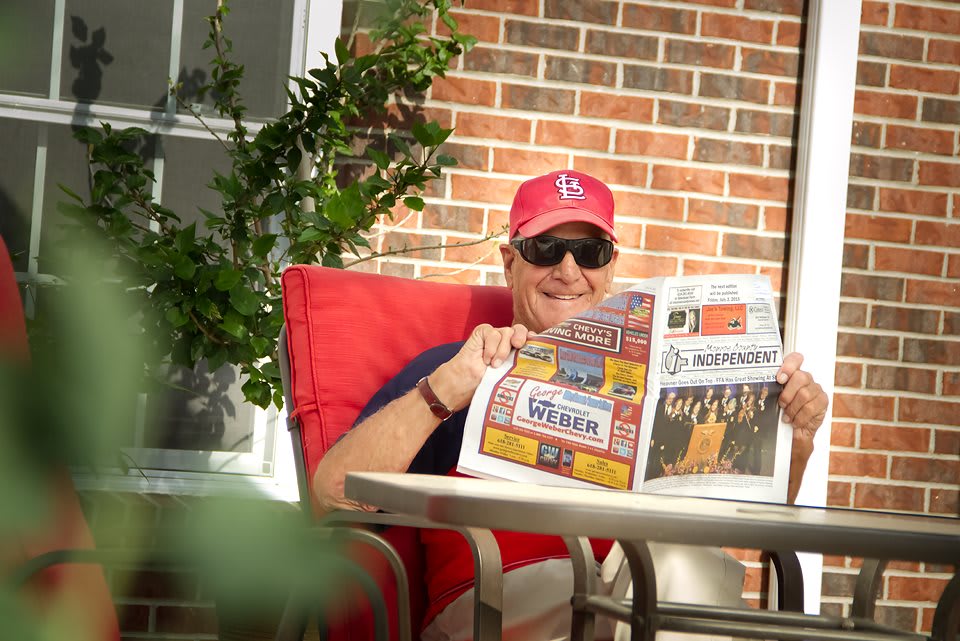 A person relaxing on a patio chair, reading a newspaper while wearing sunglasses and a cap.