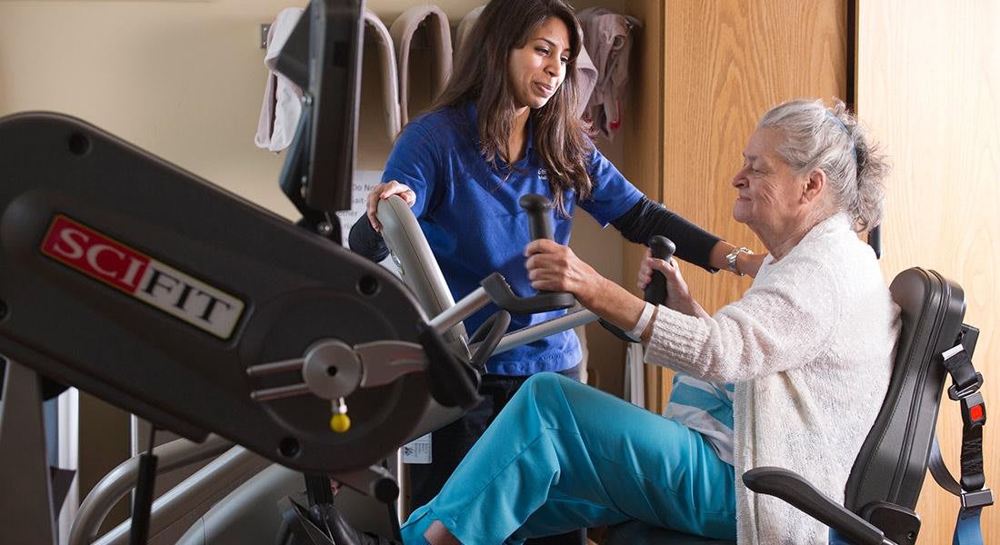 A therapist assists an elderly woman with exercise on fitness equipment.