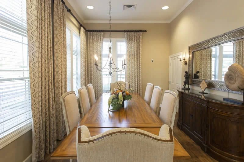 An elegant dining room featuring a long wooden table, surrounded by upholstered chairs and adorned with a central floral arrangement. Large windows with patterned curtains allow natural light to illuminate the space, complemented by a ceiling chandelier.