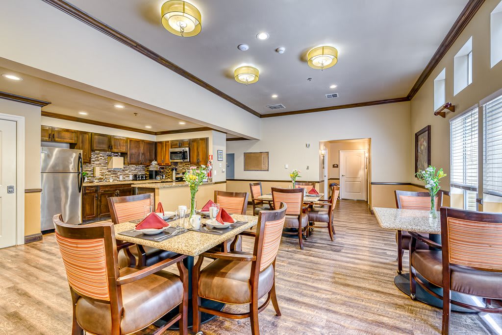 A well-lit dining area with wooden flooring, neatly arranged tables and chairs, and a view of an open kitchen.