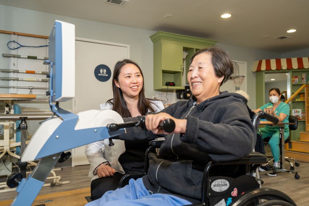 A person in a wheelchair using exercise equipment with the assistance of a healthcare professional.