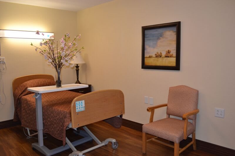 A well-furnished hospital room featuring a medical bed, chair, decorative lamp, and a wall painting.