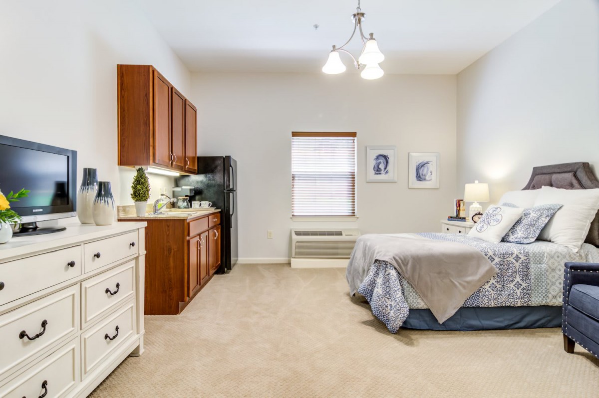 A cozy bedroom with a kitchenette, featuring wooden cabinets, a bed with light-colored linens, and soft lighting.