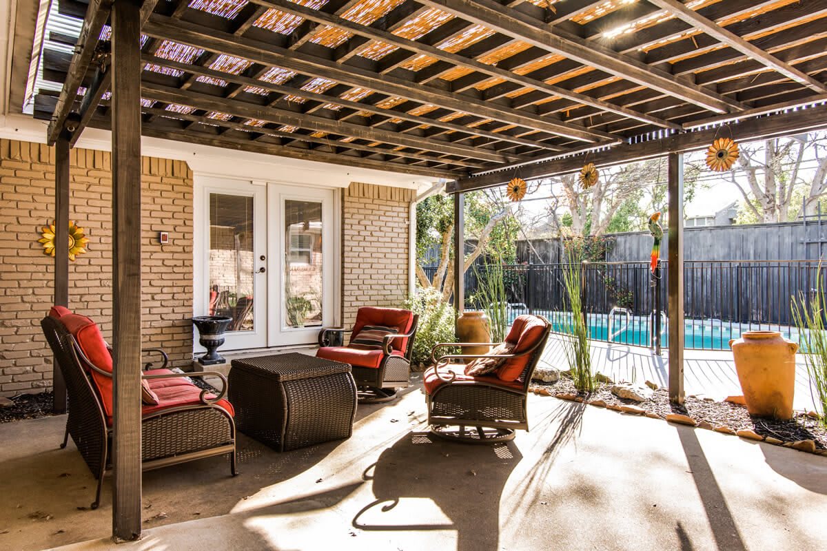 A cozy outdoor patio with comfortable seating under a pergola, overlooking a pool.