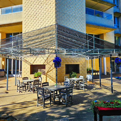 A modern outdoor cafe area featuring seating arrangements under a metal framework, surrounded by lush plants.