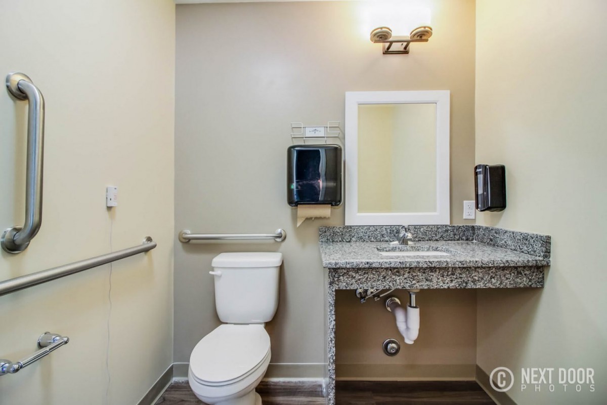 A modern bathroom with accessibility features, including a toilet, sink, mirror, and handrails.