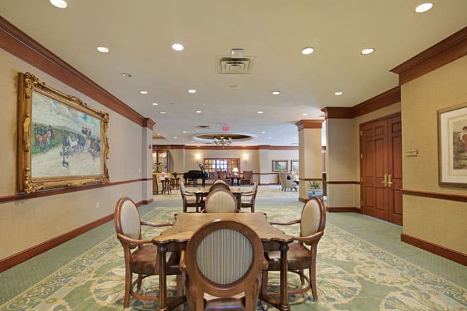 A spacious dining area with elegant furniture, featuring round tables and upholstered chairs, accented by a large painting and ceiling lights.