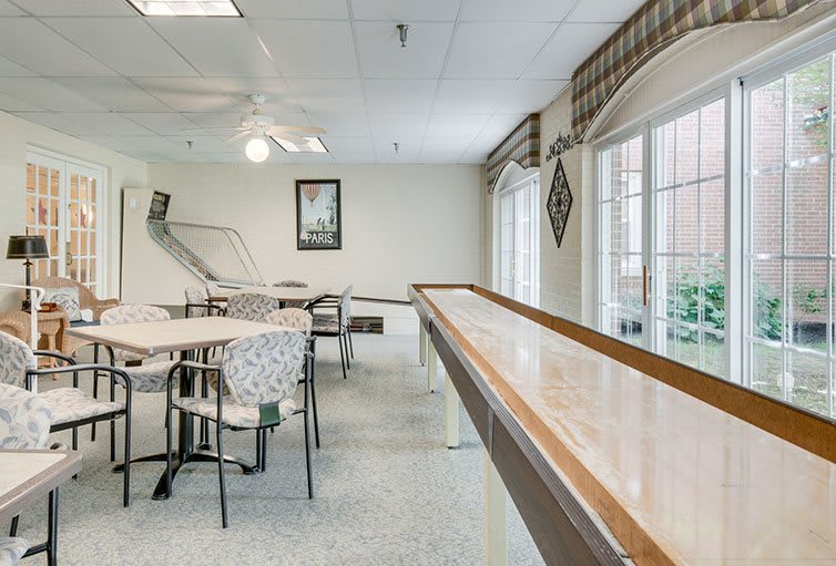 A bright and inviting game room featuring a shuffleboard table, chairs, and tables.