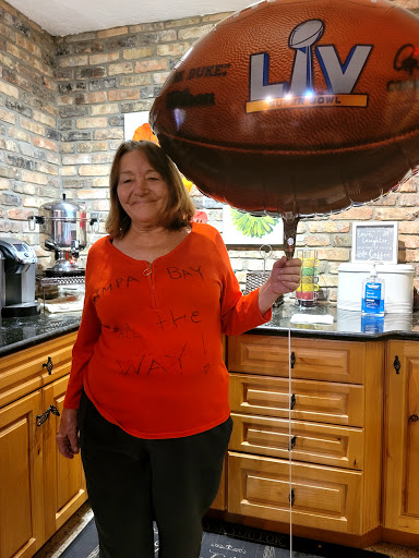 A woman in an orange shirt holding a football-themed balloon in a cozy kitchen.