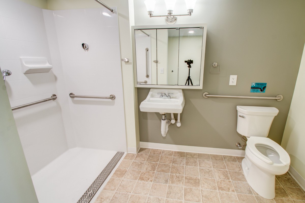 An accessible bathroom featuring a shower with grab bars, a wall-mounted sink with a mirror above, and a toilet.