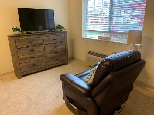 A cozy living room with an armchair facing a television on a dresser, next to a large window.
