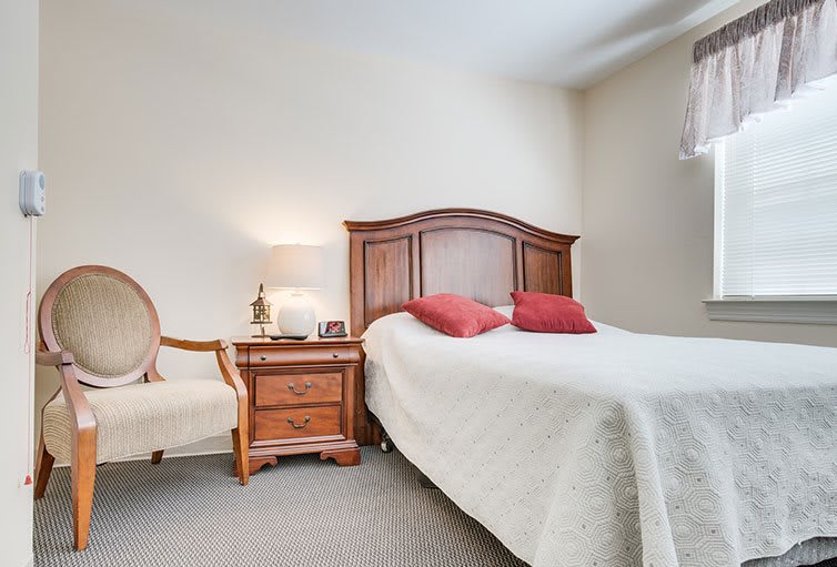 A cozy bedroom featuring a wooden bed with white bedding and red cushions, complemented by a matching nightstand and a chair.
