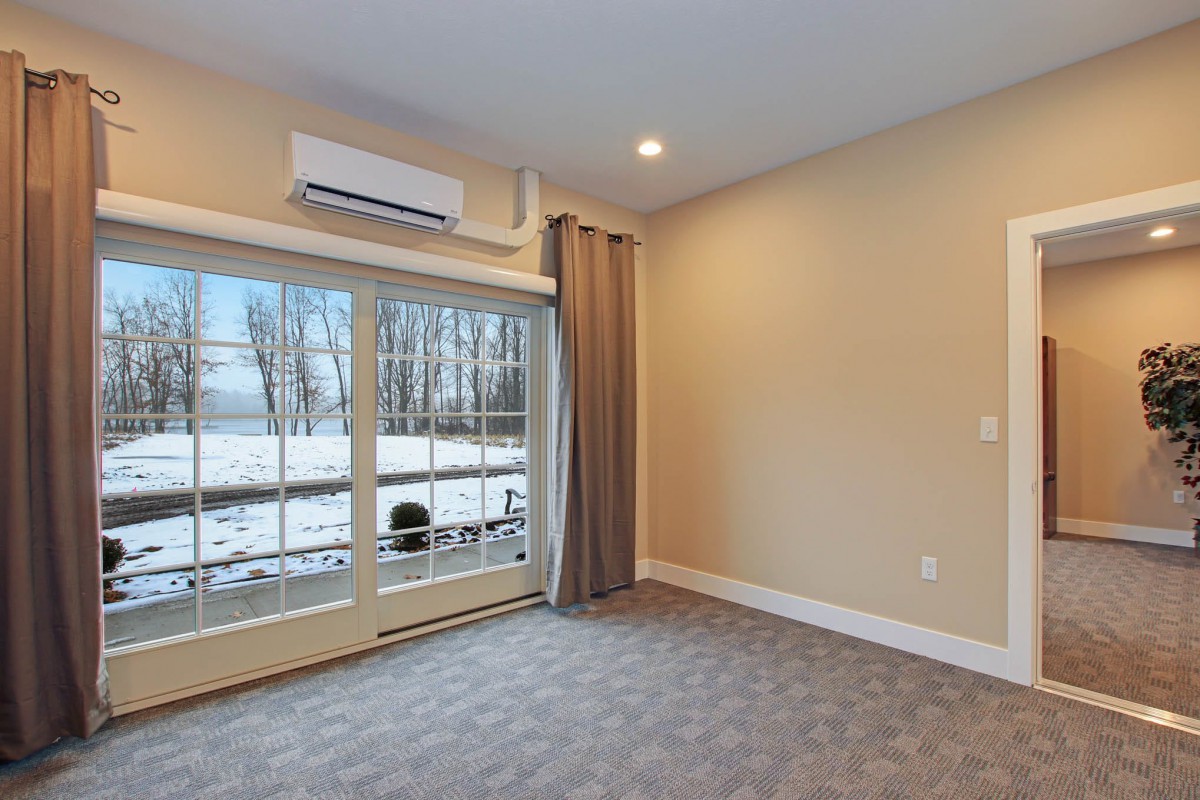 An empty room with carpeted flooring, featuring large glass doors and a winter view outside. The room is equipped with an air conditioning unit and curtains.