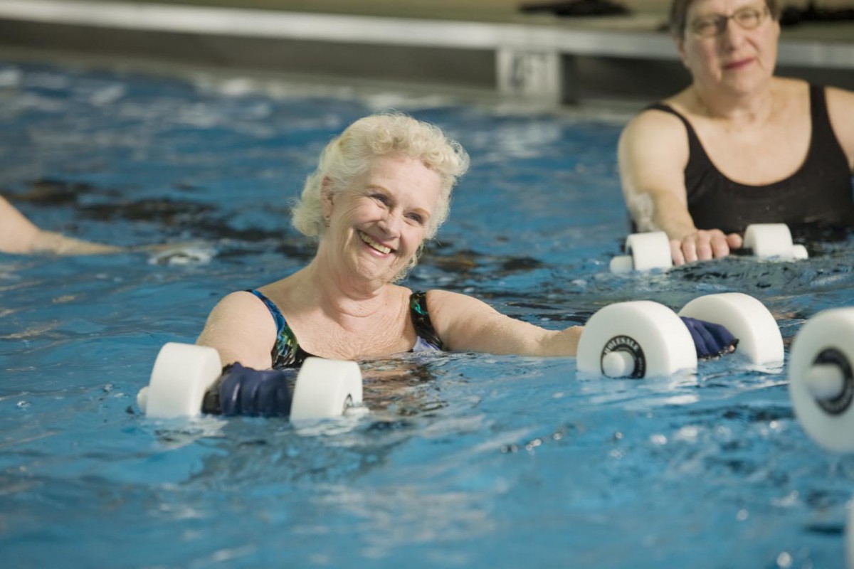 A senior woman participates in a water aerobics class, using foam dumbbells in a swimming pool.