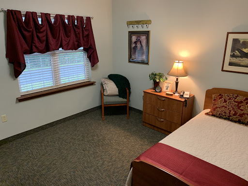 A cozy bedroom featuring a bed, a wooden dresser, a lamp, and a chair by the window.