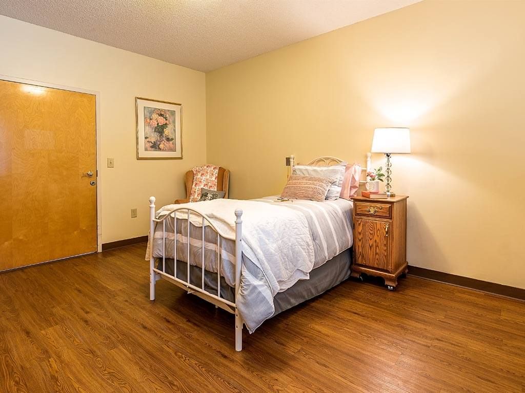 A cozy bedroom with a bed, nightstand, lamp, and chair on a wooden floor.