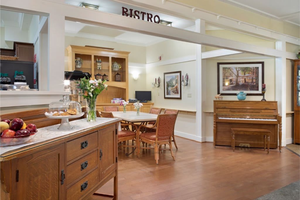 A cozy bistro interior featuring a dining area with wooden furniture, a piano, and decorative flowers.