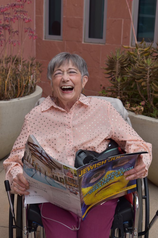 An elderly woman is smiling while reading a magazine outdoors.