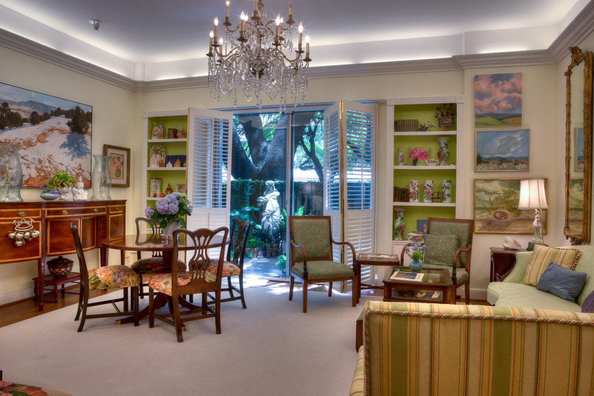 A beautifully decorated living room with traditional furnishings, a chandelier, and large windows opening to a garden view.