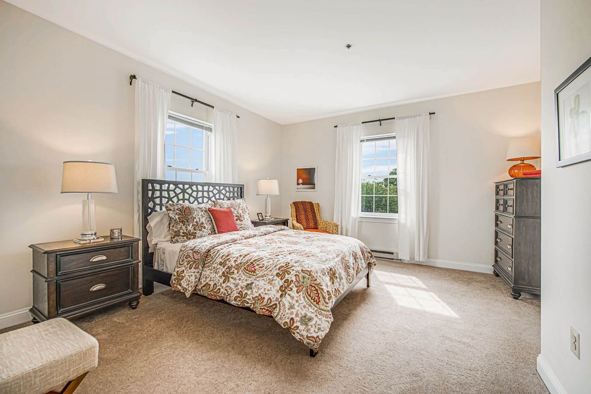A cozy bedroom featuring a patterned bedspread, two windows with curtains, and warm lighting.