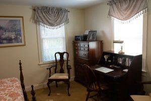 A cozy home office with vintage furniture, featuring a desk and chair near two windows.