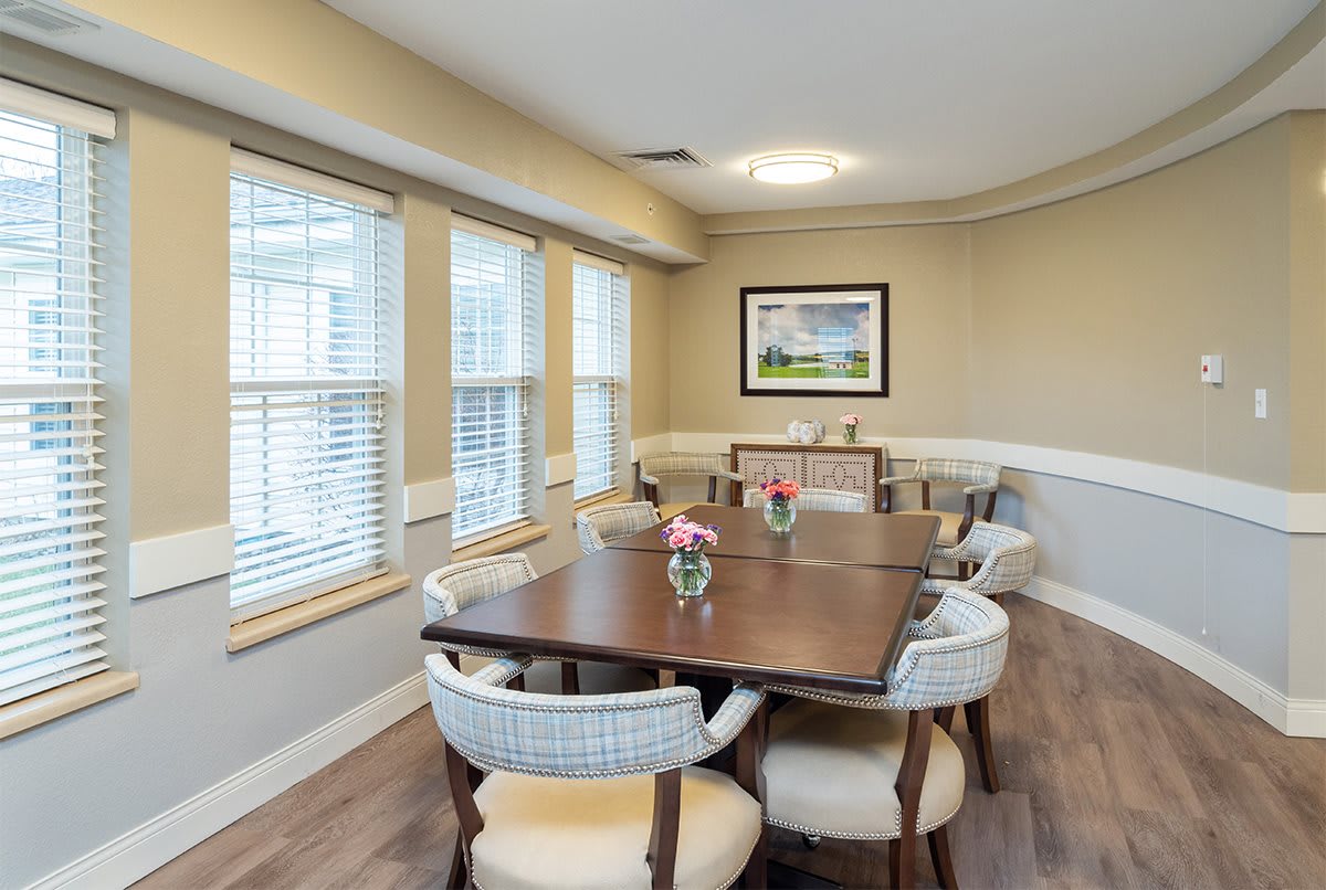 A cozy dining room with a large wooden table and upholstered chairs, accented by flowers and natural light streaming through multiple windows.