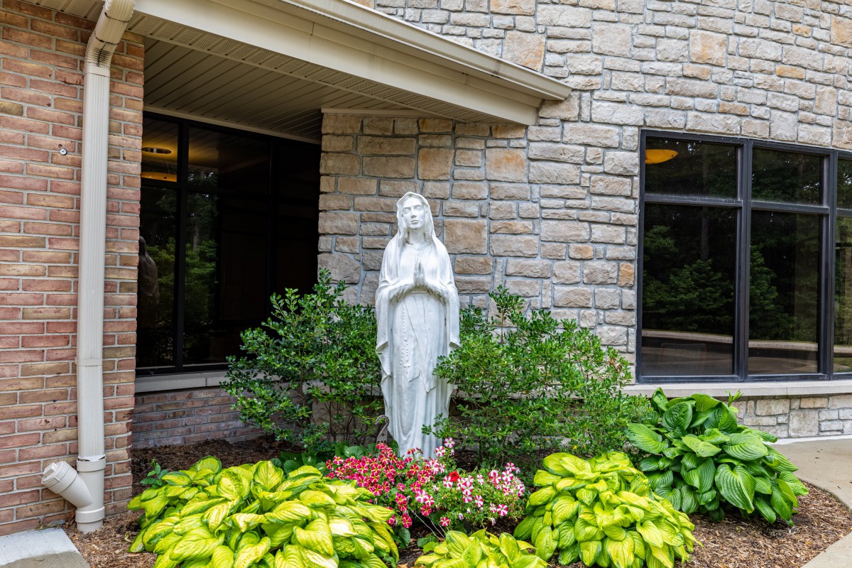 A religious statue surrounded by lush greenery in a garden setting beside a building.