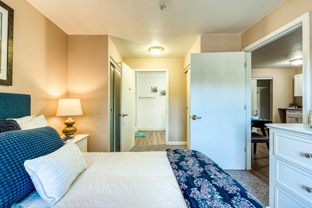 A well-lit bedroom featuring a neatly made bed with patterned bedding, a bedside lamp, and an open doorway leading to another room.