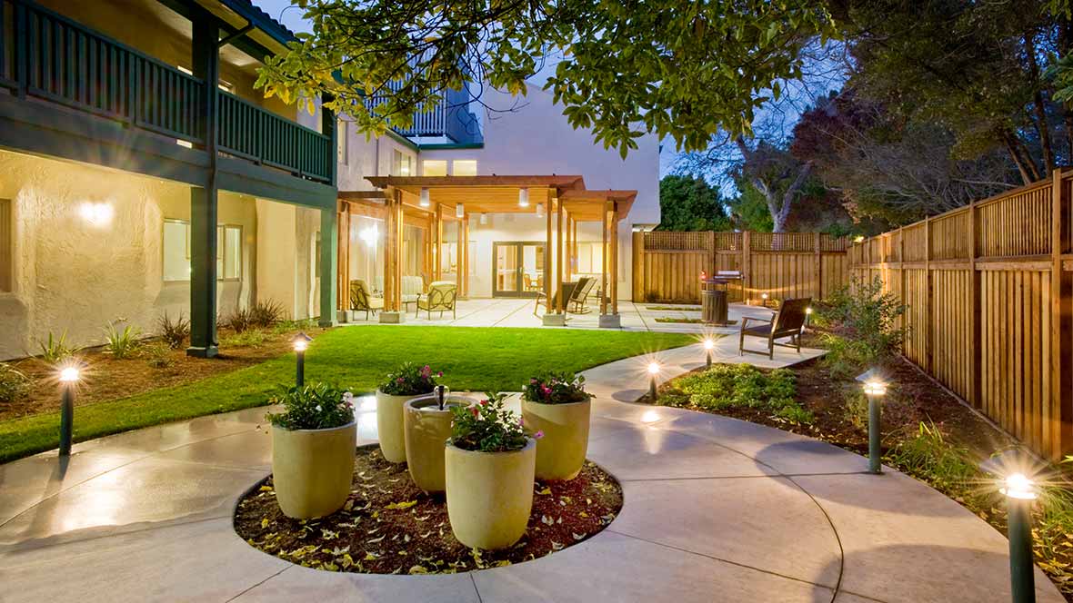 A beautifully landscaped backyard with a patio and garden, illuminated by evening lights.